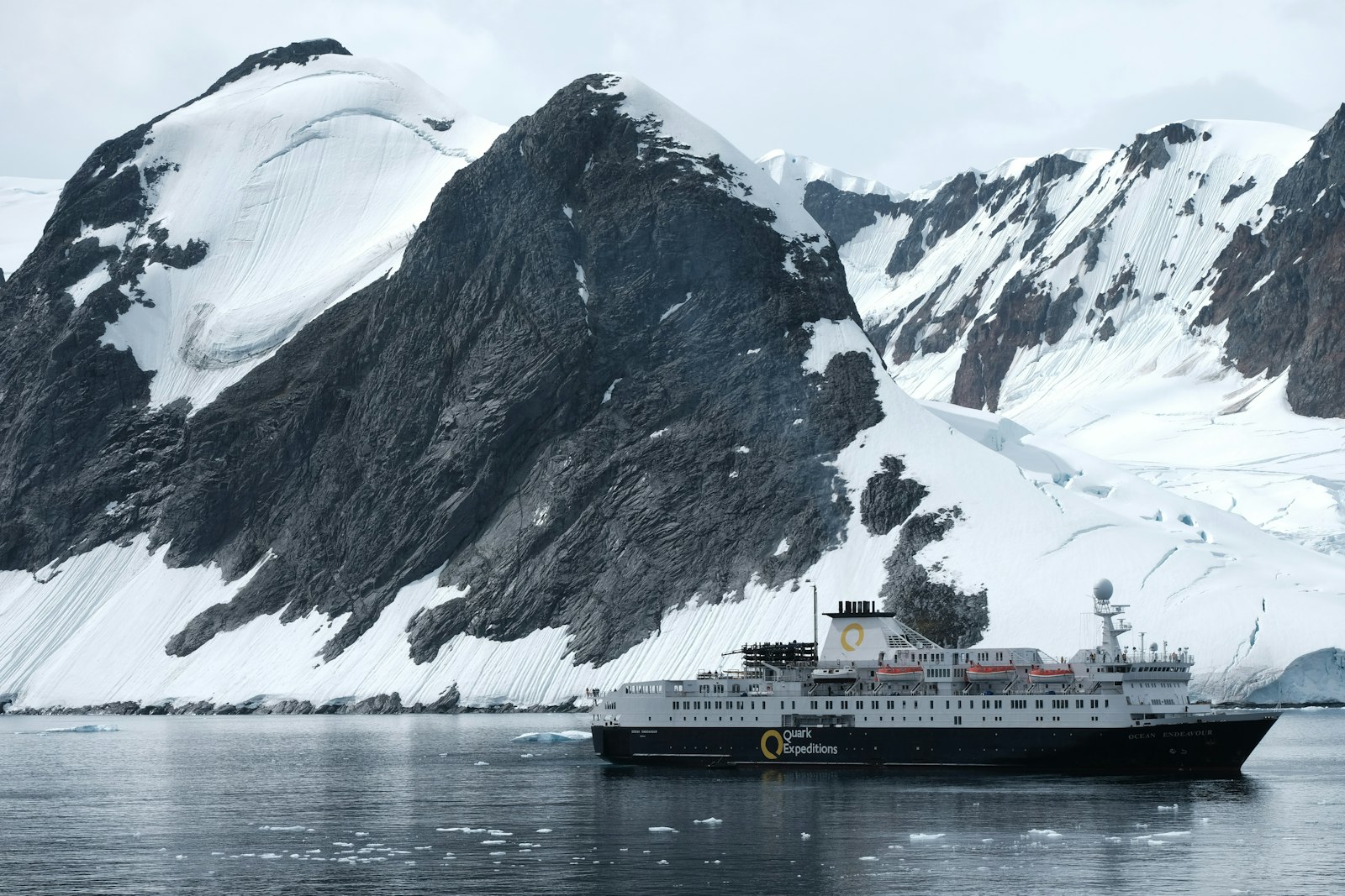 A polar expedition cruise ship sailing beneath dramatic snow-covered Antarctic peaks